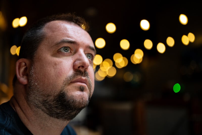 A man with short dark hair and a beard is looking slightly upwards with a contemplative expression. The background is out of focus, with warm, circular bokeh lights creating a cozy atmosphere.