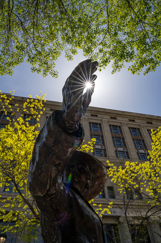 A bronze statue of a person shading their eyes with a raised hand stands amidst a backdrop of a building and green trees. The sun peeks through the statue's fingers, creating a starburst effect.