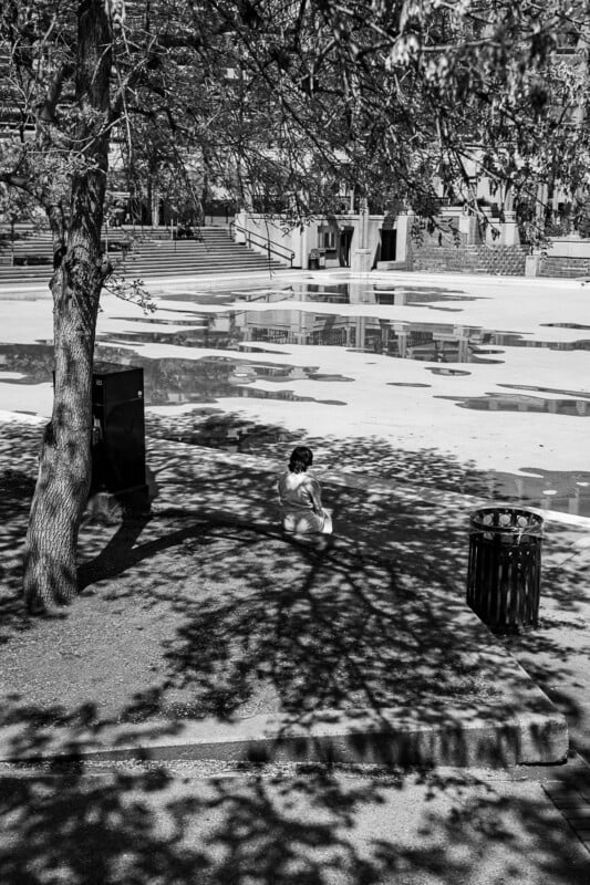 Black and white image of a child sitting alone on a sunlit pavement near a tree in a large, empty, urban square. The tree and child create distinct shadows on the ground. A trash bin is nearby and buildings are visible in the background.