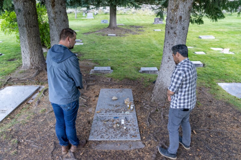 Two men standing by a grave in a cemetery, surrounded by trees. one man is observing the grave while the other appears reflective. grave markers can be seen in the background.