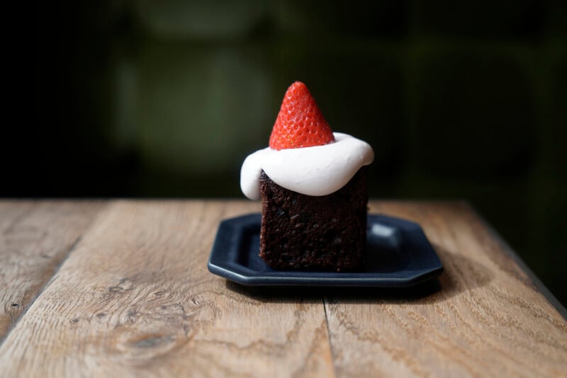 A slice of chocolate cake with white frosting and a halved strawberry on top sits on a small, dark square plate. The plate rests on a wooden table with a blurred green background.