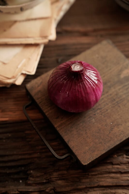 A whole red onion sits on a rustic wooden cutting board with a metal handle. The background features a stack of aged papers and a wooden surface, contributing to a vintage and homely atmosphere.
