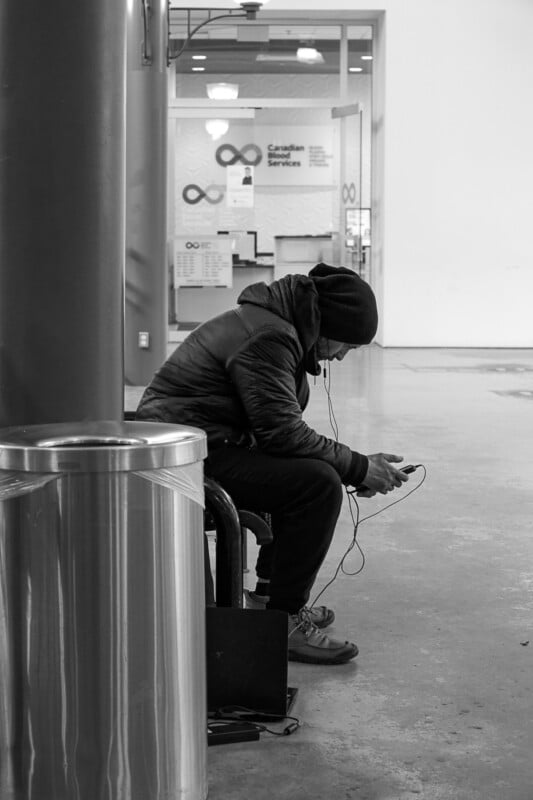 A person wearing a hoodie and jacket sits on a bench, hunched forward, using a mobile device with earphones in. A metal trash can is in the foreground. Behind, a door with "Canadian Services" and infinity symbols is partially visible. The scene is indoors.