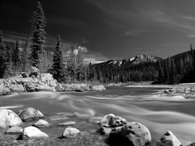 Black and white image of a serene river flowing through a rocky landscape with pine trees and mountains in the background under a clear sky.