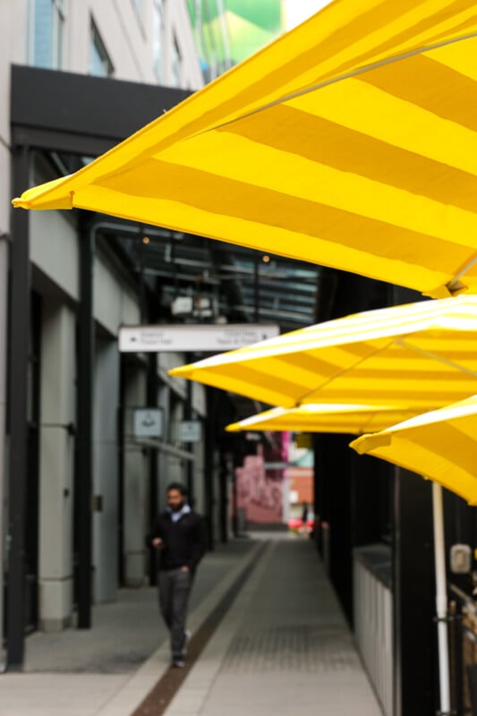 A series of bright yellow umbrellas lines a narrow alleyway. In the background, an individual walks along the path, with modern buildings on either side. Directional signs hang above the alley, adding an urban touch to the scene.