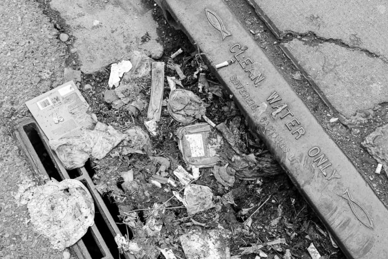 Black and white image of litter that includes a cigarette pack and other trash collected alongside a storm drain marked "C. GEN. WATER ONLY.