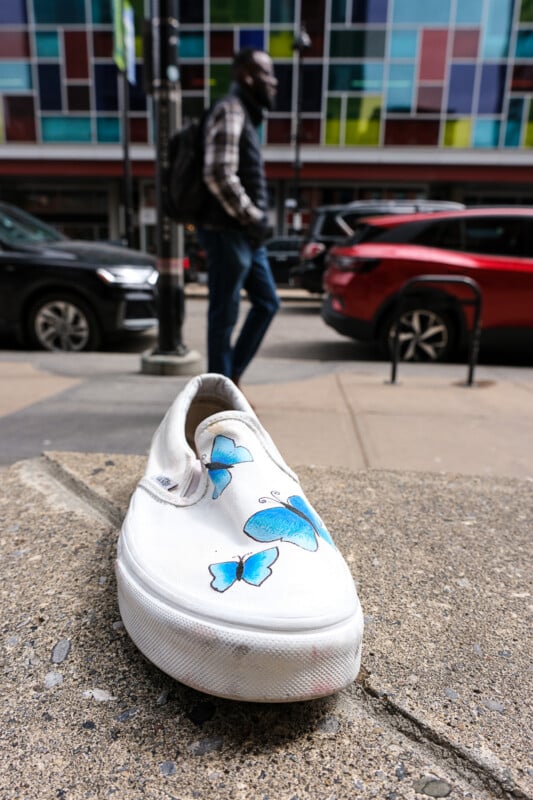 A white slip-on shoe with blue butterfly designs on its top sits on a concrete ledge, with parked cars and a person walking by in the background. The scene is set in an urban area, featuring a colorful glass-paneled building.