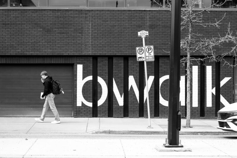 A person wearing a backpack walks past a brick building with large, partially visible white letters on it. Two parking signs are mounted on a pole in the foreground, and the sidewalk has a small tree with bare branches. A car is partially visible on the right.