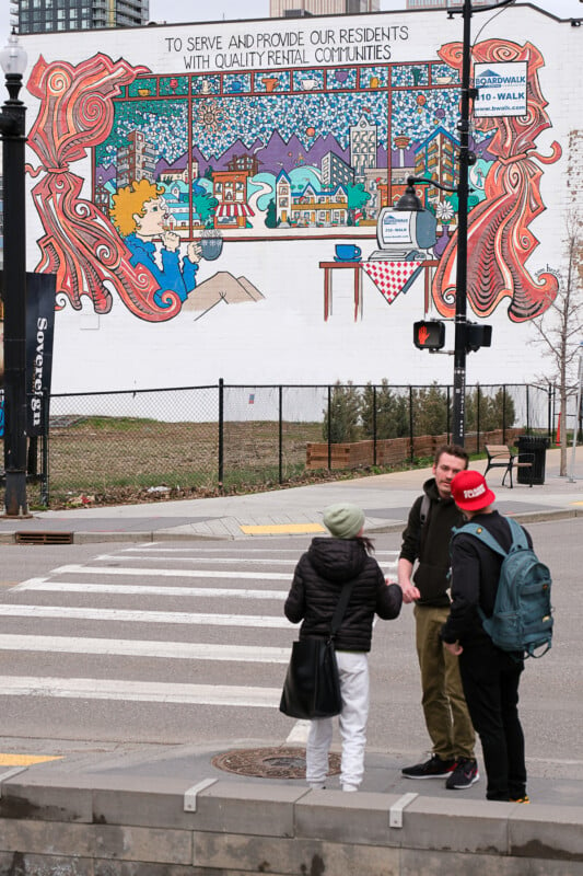 Three people stand at a crosswalk, engaging in conversation, against a backdrop of a vibrant, colorful mural painted on a building. The scene captures urban life and street art.
