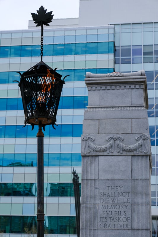 A close-up photo of a memorial flame in front of a stone monument inscribed with the words "THEY SHALL NOT DIE WHILE MEMORY FULFILS ITS TASK OF GRATITUDE." A modern glass building with teal and blue windows is visible in the background.