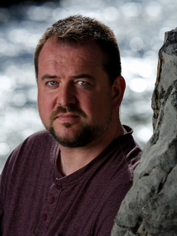A man with a beard and short hair, wearing a maroon shirt, looks intently at the camera. He stands near a rock with a blurred silver background.