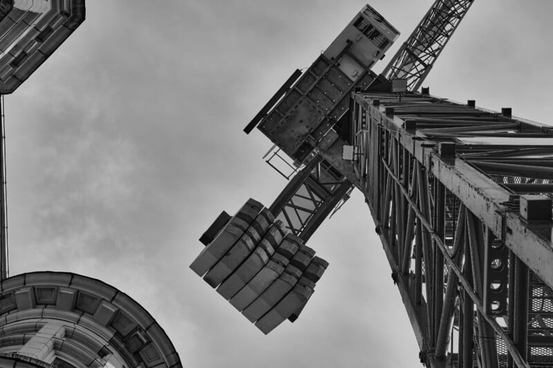 Black and white photograph of a high-rise construction crane, viewed from below, lifting building materials. The crane's metal structure contrasts against a cloudy sky. Part of a nearby building is visible on the left edge of the image.