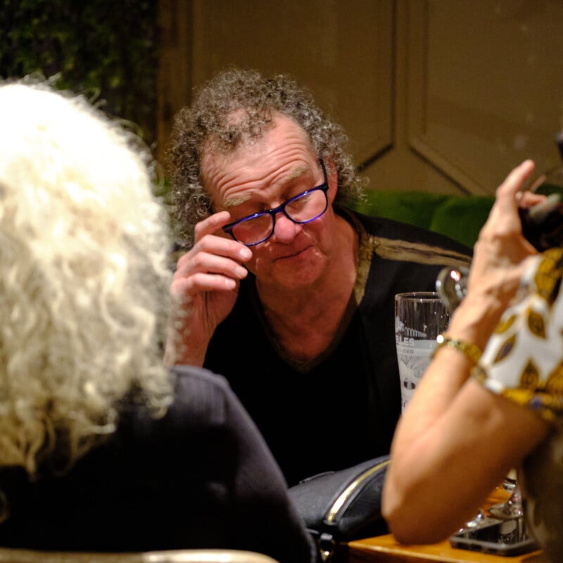A man with curly hair and glasses sits at a table, appearing to adjust his glasses and engaging in conversation with others. He is surrounded by two people, one with white hair and the other with short hair. The table holds a glass and some items.