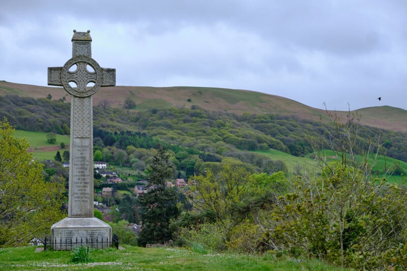 A stone Celtic cross gravestone stands in a green, hilly landscape with small houses and trees in the background. The sky is overcast, casting a soft light over the scenery. A single bird is flying near the right side of the cross.