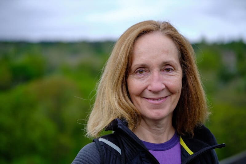 A woman with shoulder-length light brown hair is smiling at the camera. She is wearing a black jacket with a purple shirt underneath. The background is an outdoor setting with green trees and an overcast sky.