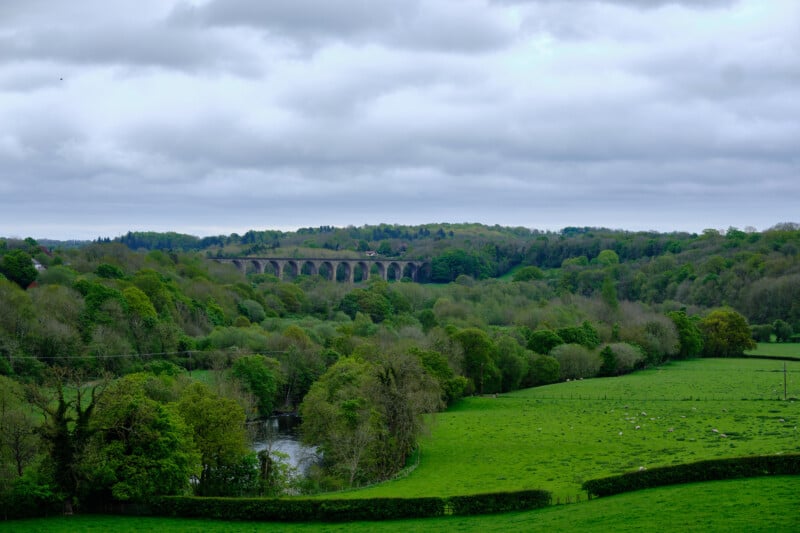 A lush green landscape with rolling hills and dense woods. A viaduct with arched supports extends across the scene in the distance under a cloudy sky. A calm river winds through the fields in the foreground. Scattered sheep graze in the fields.