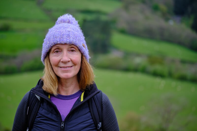A woman with shoulder-length blonde hair is smiling while wearing a purple knitted hat, a purple top, and a black jacket. She has a backpack on and greenery with trees and fields is visible in the blurred background.