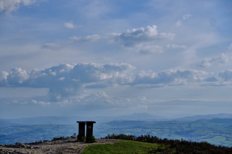 A scenic image of a mountain summit under a vast blue sky with fluffy clouds. A stone trig point stands on the peak, overlooking a sprawling, verdant landscape of rolling hills and valleys.