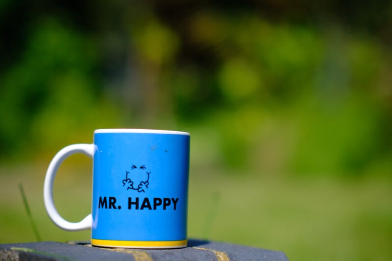 A blue coffee mug with "MR. HAPPY" printed in black text and a simple cartoon face sits on a flat surface. The background is blurred with green foliage, suggesting an outdoor setting.