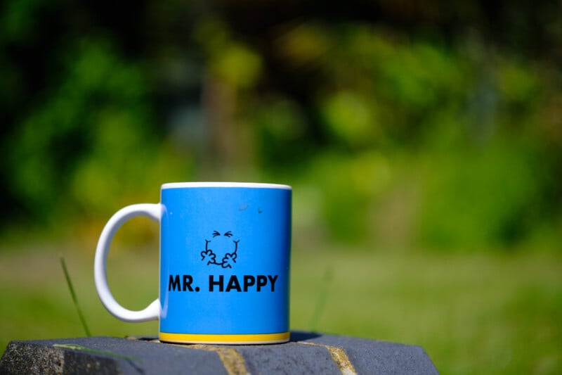 A blue mug with the text "MR. HAPPY" and a small cartoon face is placed on a stone surface. The background features a lush, green, out-of-focus garden or outdoor space, creating a vibrant and cheerful atmosphere.