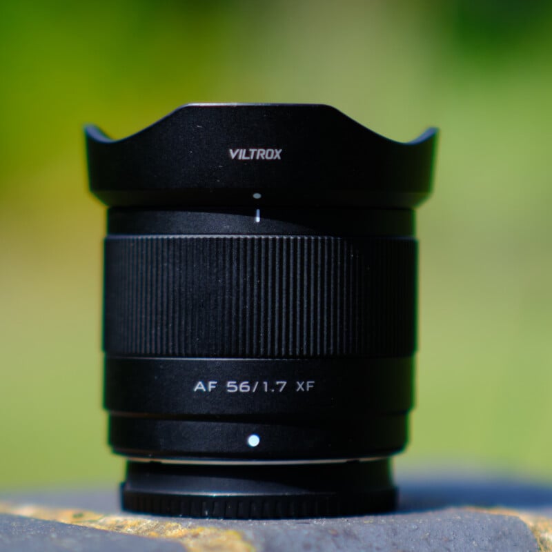 A close-up of a black Viltrox AF 56mm f/1.7 XF camera lens, with fine details visible in its design and texture. The lens is positioned against a blurred, natural green and yellow background, highlighting its sleek and professional appearance.