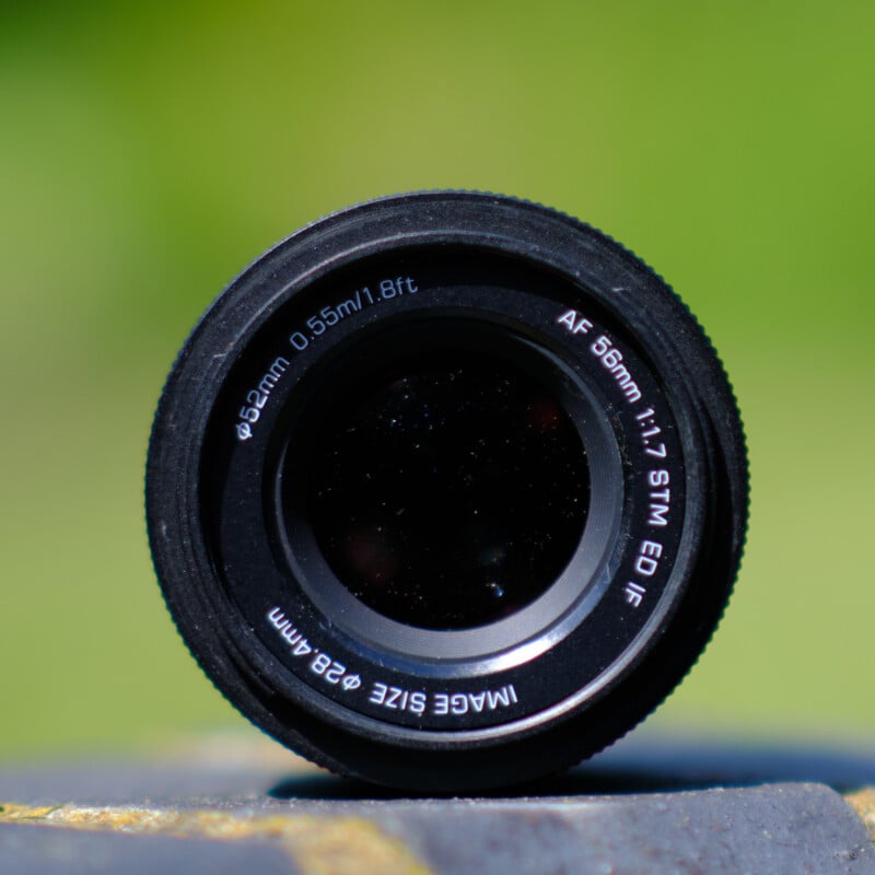 Close-up photograph of a camera lens with specifications: AF 56mm 1:1.7 STM ED IF, lens size Ø 62mm, and focusing distance 0.55m/1.8ft. The background is blurred, with shades of green and yellow indicating an outdoor setting.