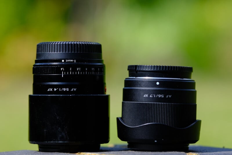 Close-up of two camera lenses standing upright on a flat surface outdoors. The lens on the left is taller with a focus ring and labeled "AF 56/1.4 XF," while the shorter lens on the right is labeled "AF 18/2 XF." Both lenses have a black finish.