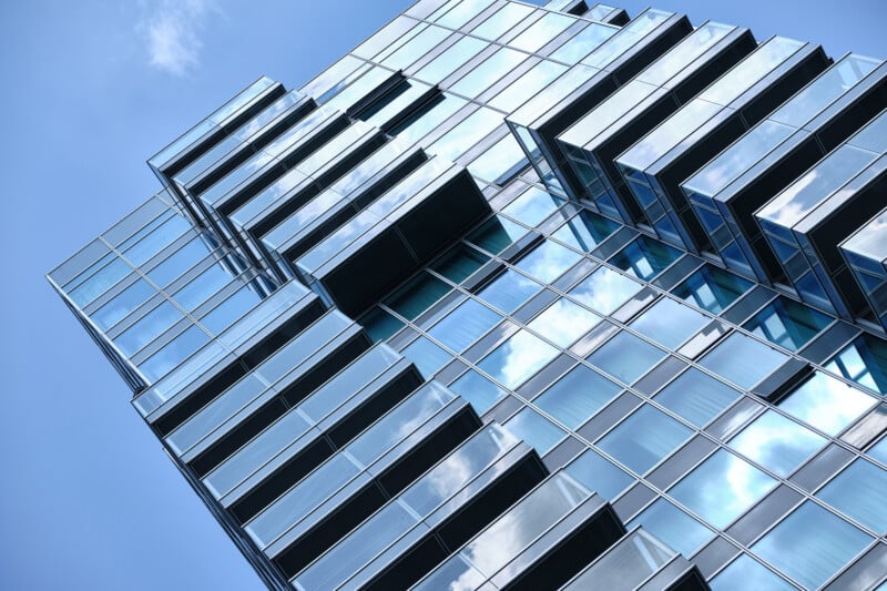A modern high-rise building with a reflective glass facade against a clear blue sky. The building features staggered sections and balconies, creating a geometric pattern. The glass reflects the sky and clouds.