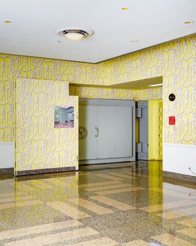 A room with bright yellow patterned wallpaper and a shiny, patterned floor. There is a large, closed, grey, metal door in the center, a red fire alarm on the right wall, and an image of a building on the left wall. A circular light fixture is on the ceiling.