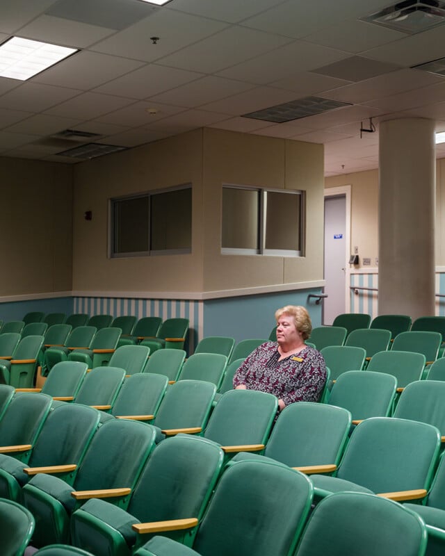 A woman sits alone in an empty auditorium with rows of green chairs. She is wearing a patterned top and appears to be waiting or observing something. The room has beige walls with small windows and a door in the background. The scene is well-lit with overhead lights.