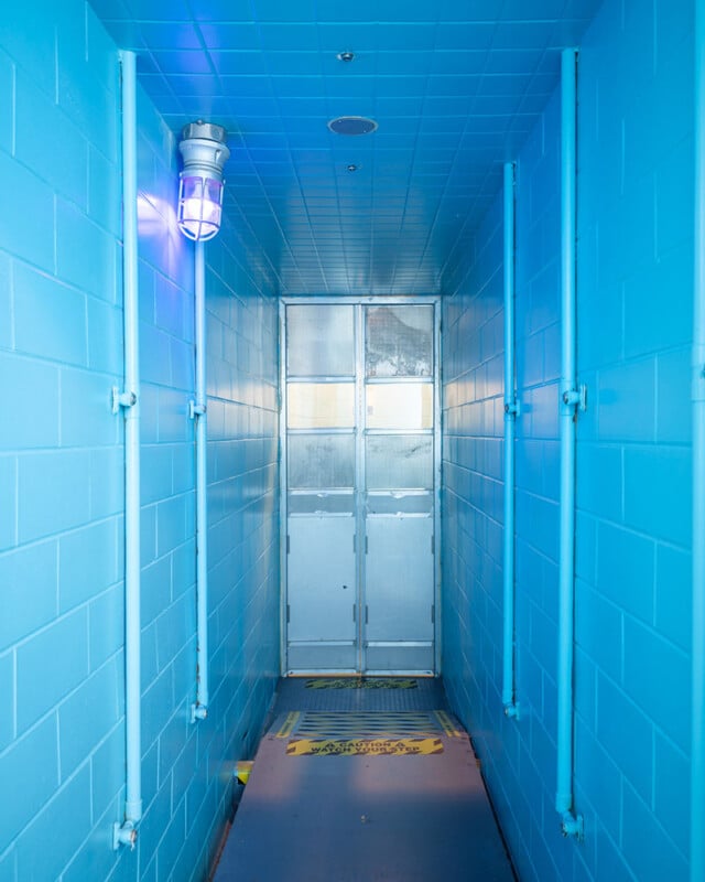 A narrow hallway with blue-tiled walls and ceiling, illuminated by a blue-tinted light fixture on the left wall. The hallway leads to a set of double doors at the end. Caution signs are visible on the floor.