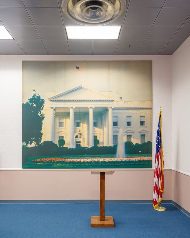 A small podium stands in front of a large photograph of the White House, displayed on a wall in a room with blue carpeting. An American flag is placed to the right of the podium. The ceiling features a circular vent.