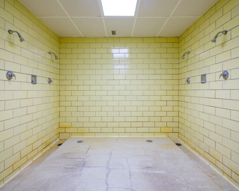 A spacious communal shower room with cream-colored tiled walls and multiple showerheads arranged on the side walls. The floor is covered with matching tiles, and the room is brightly lit by overhead fluorescent lights.