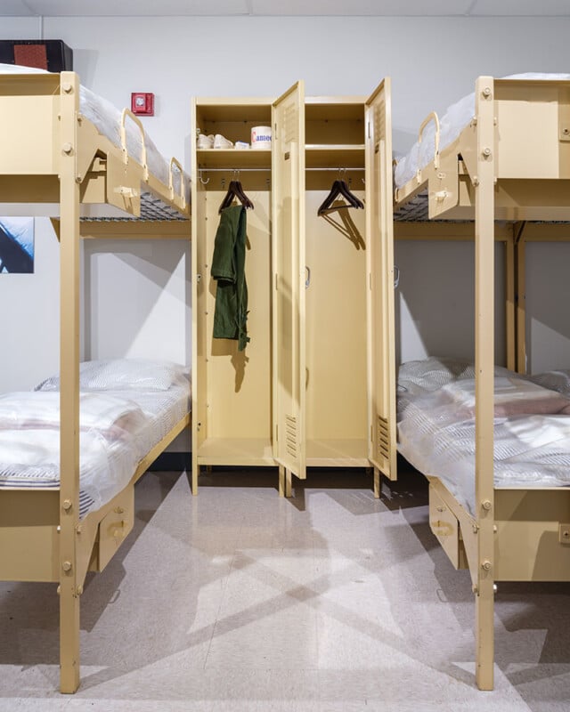 A tidy military-style dorm room with two sets of tan metal bunk beds, one on each side. The center has an open locker displaying a green uniform, hangers, and a small canister on the top shelf. The room has overhead lighting and a shiny floor.