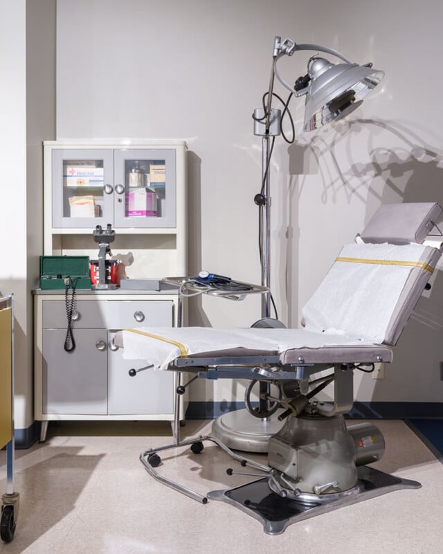 A medical examination room with an adjustable chair, covered with white paper, situated under a large examination lamp. A medical supply cabinet containing various items is in the background, along with a blood pressure monitor and other medical instruments.