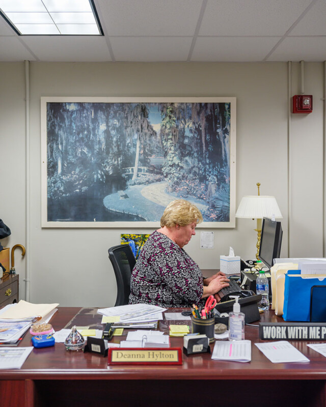 A person with short blond hair sits at a desk typing on a computer keyboard. The desk is cluttered with various office supplies and papers, and a nameplate reads "Deanna Hylton." Behind them is a large landscape painting, and the room is well-lit with overhead lights.