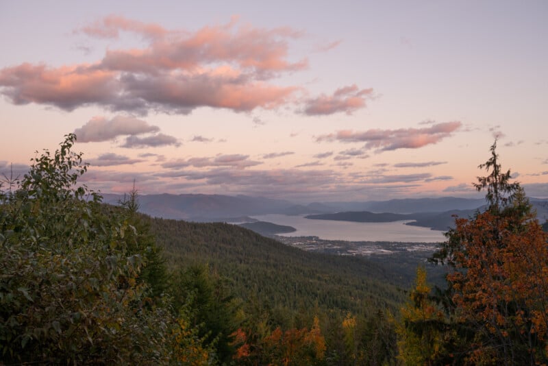 A panoramic view of a lake surrounded by autumn-colored forests and mountains at sunset, with pink clouds strewn across the sky.
