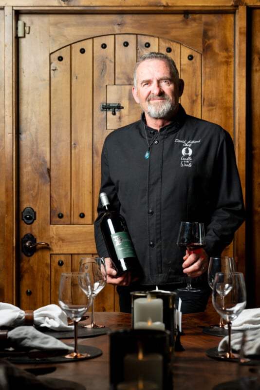 A distinguished older man with a beard, dressed in a black chef jacket, holding a bottle of wine and a wine glass, standing in front of a wooden door in a rustic dining setting.