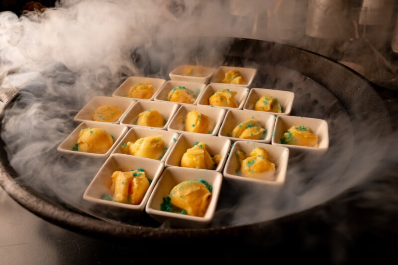 A circular tray of bite-sized appetizers in small white dishes, shrouded in dramatic culinary smoke, creating an enticing and elegant presentation.