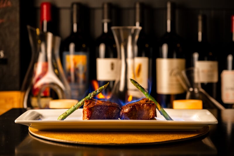 A gourmet plate with two succulent steak medallions and asparagus, elegantly presented on a long dish against a background of wine bottles. the setting is warmly lit, creating an inviting dining ambiance.