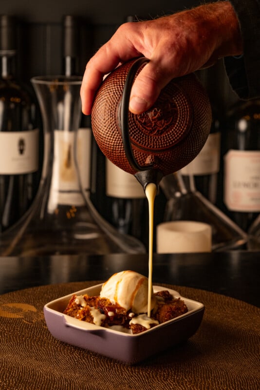 A person pours a thick, golden syrup from a small, textured ceramic jug over a scoop of vanilla ice cream served on a bed of crumbled dessert, with wine bottles blurred in the background.