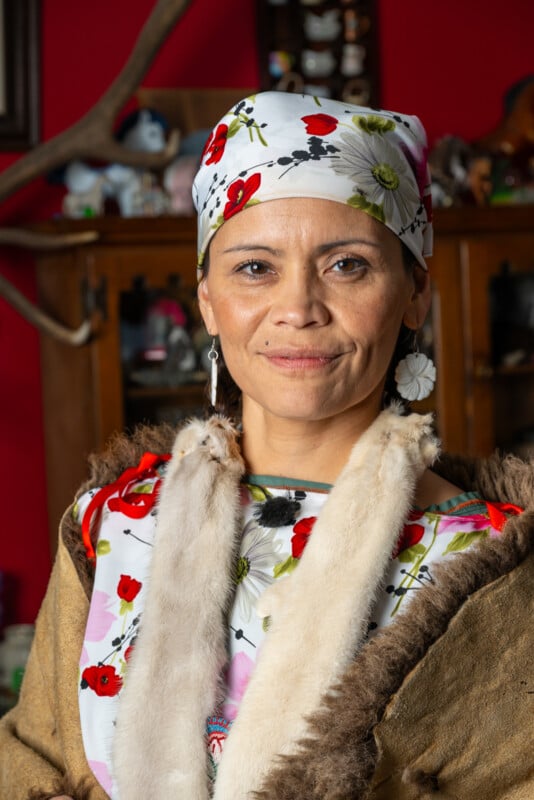 A woman wearing a colorful headscarf and a traditional fur-trimmed outfit stands in front of a bookshelf, smiling at the camera.