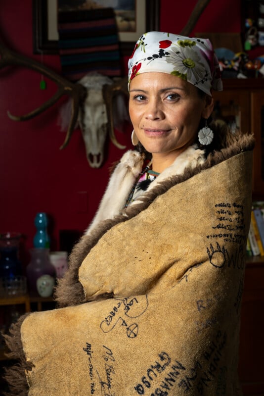 A woman with warm eyes, wrapped in a hand-signed fur-lined cloak, stands confidently in a colorful room decorated with eclectic items, including a mounted goat head.