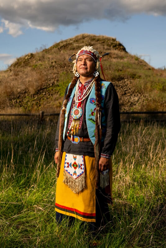 An indigenous person stands outdoors in traditional regalia including a beaded headband, braided hair, a fringed vest, and a brightly colored, beaded long skirt, with the backdrop of a grassy field and small hill under a cloudy sky.