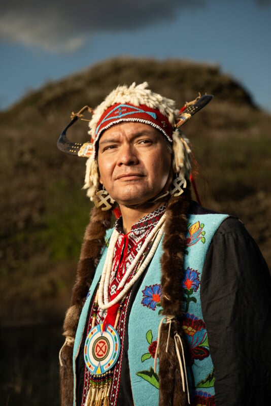 A native american man in traditional attire, including a feathered headdress, stands proudly outdoors with a rocky hill in the background during sunset.