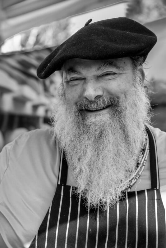 Black and white portrait of a cheerful elderly man with a long white beard, wearing a beret and striped apron, smiling broadly at a market.
