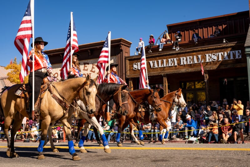 Horseback riders dressed in western attire carrying american flags at a parade in a town, with spectators watching from the sides under a clear blue sky.