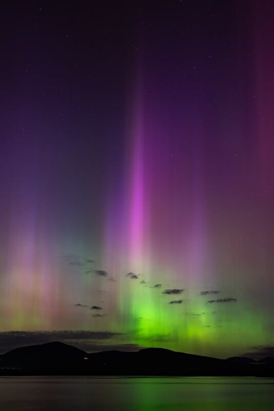 Vibrant northern lights (aurora borealis) display vivid purple, green, and pink streaks extending upwards over a shadowy lake and mountain horizon under a starlit sky.