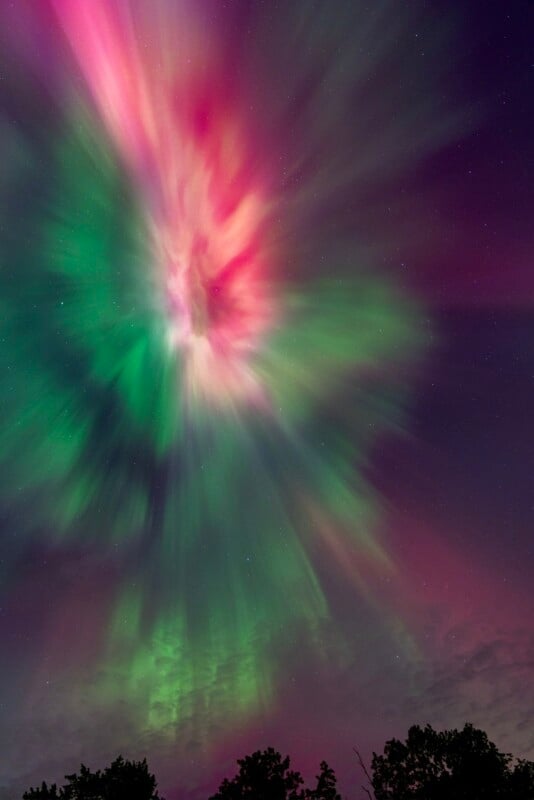 A vibrant aurora borealis displays bursts of pink and green light spreading across a night sky, with silhouetted treetops visible at the bottom.