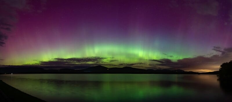 Panoramic view of vibrant Aurora Borealis reflecting on a serene lake with distant mountains under a starry night sky.